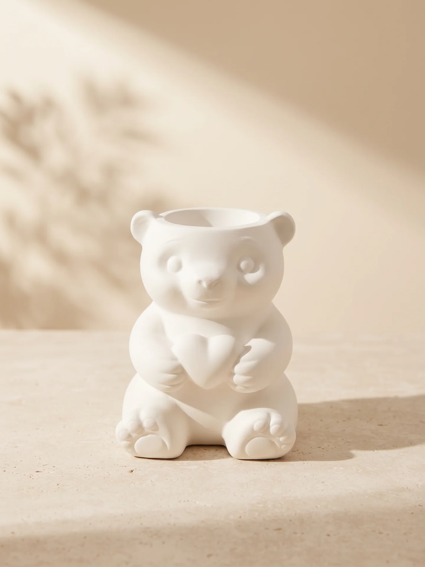 Portalápices decorativo de yeso blanco con diseño de osito abrazando un corazón sobre superficie de piedra caliza en un ambiente de estudio minimalista.